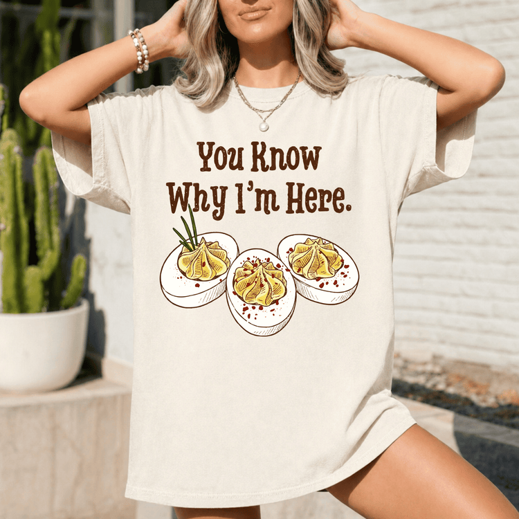 A woman wearing a white t-shirt with a graphic design of three deviled eggs and the text "You Know Why I'm Here."
