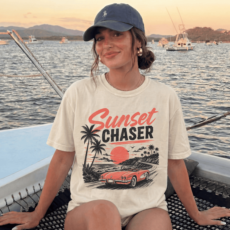 A woman wearing a white t-shirt with a graphic of a car and palm trees, sitting on a boat in the ocean at sunset.