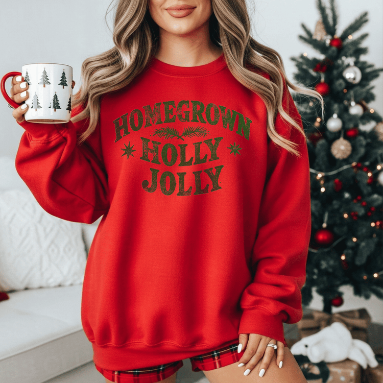 A woman wearing a red sweater with the text "Homegrown Holly Jolly" is holding a mug in front of a Christmas tree.
