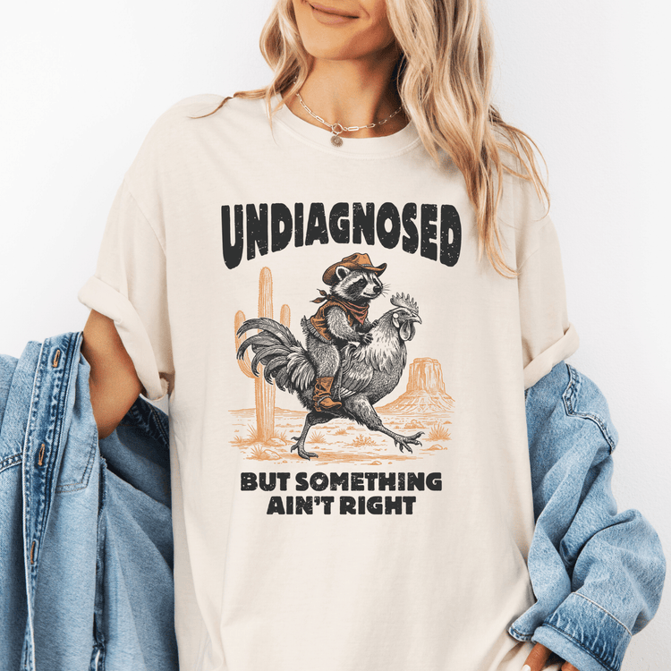 A woman wearing a white t-shirt with a graphic design of a raccoon riding a rooster and the text "UNDIAGNOSED BUT SOMETHING AIN'T RIGHT" printed on it.