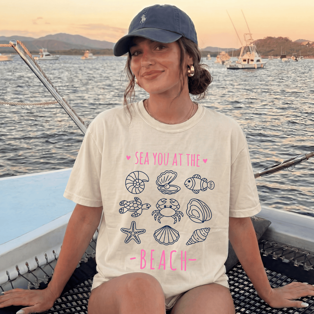 A woman wearing a white t-shirt with a graphic design of sea creatures and the text "SEA YOU AT THE BEACH" is sitting on a boat in the ocean.