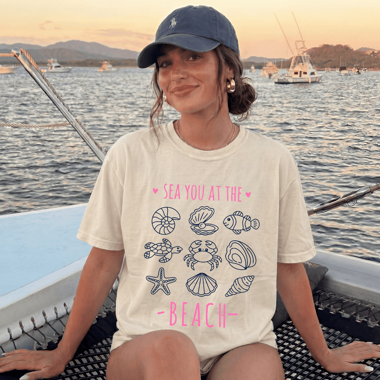 A woman wearing a white t-shirt with a graphic design of sea creatures and the text "SEA YOU AT THE BEACH" is sitting on a boat in the ocean.