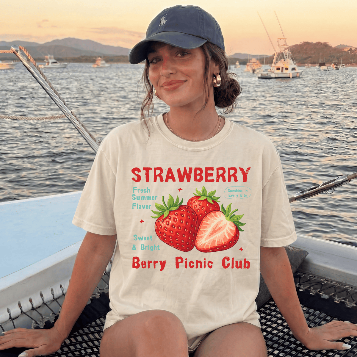 A woman wearing a white t-shirt with a strawberry graphic and text, sitting on a boat with a scenic background of mountains and boats.