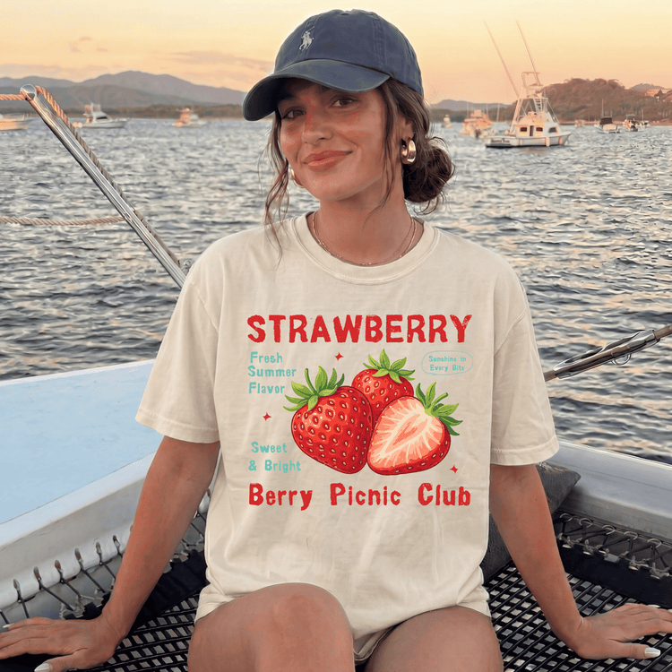 A woman wearing a white t-shirt with a strawberry graphic and text, sitting on a boat with a scenic background of mountains and boats.