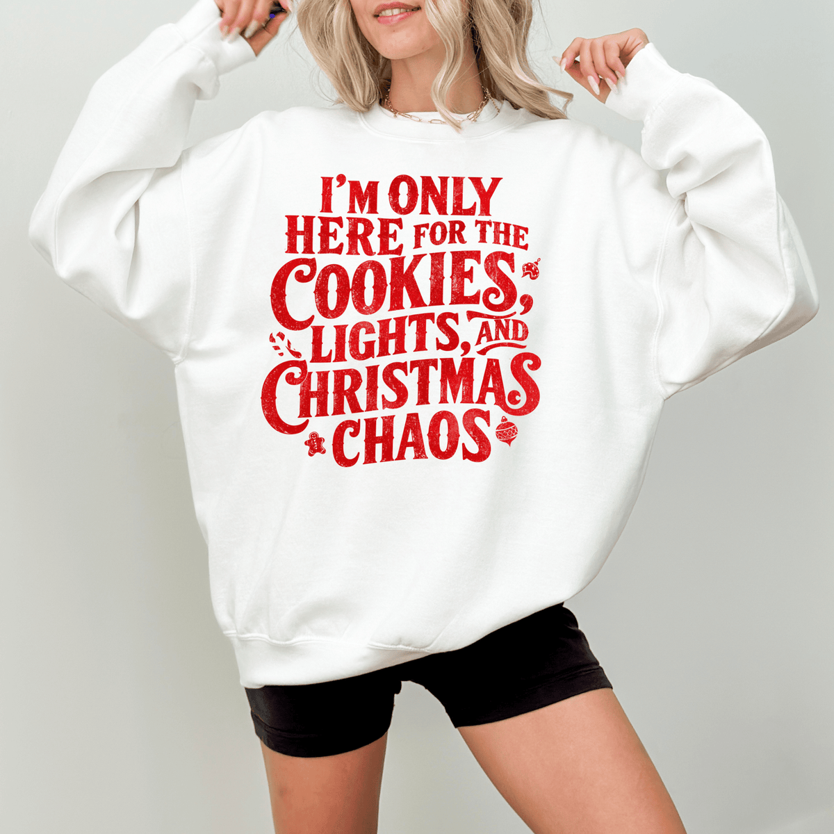 A woman wearing a white sweatshirt with a red text design that says "I'm only here for the cookies, lights, and Christmas chaos".