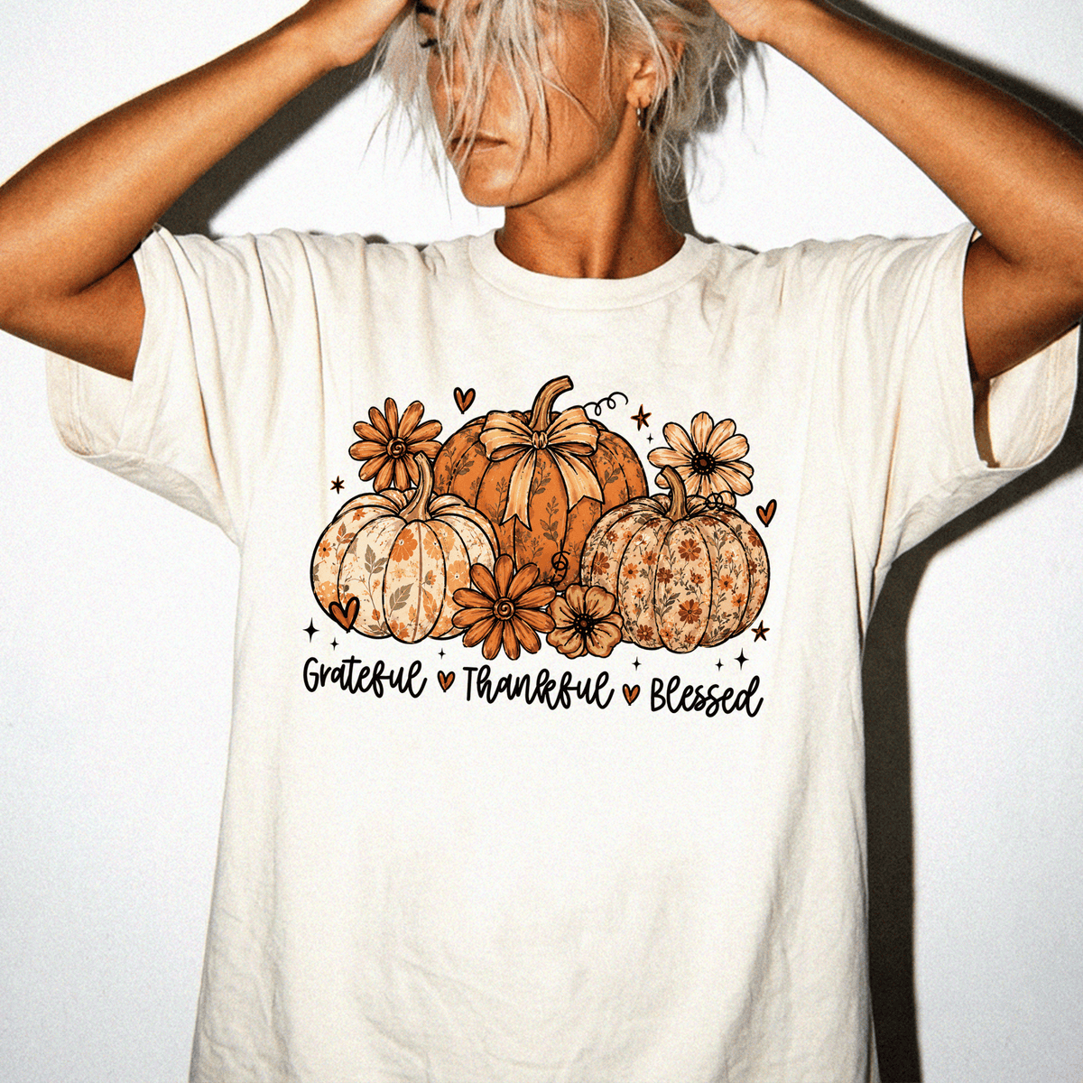 A woman wearing a white t-shirt with a graphic design of pumpkins and flowers, along with the text "Grateful * Thankful * Blessed" printed on it.