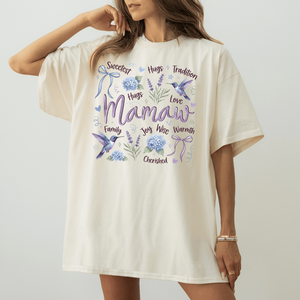 A woman wearing a white t-shirt with a graphic design featuring the word "Mamaw" surrounded by various floral and bird-related elements.