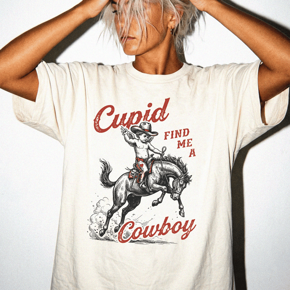 A woman wearing a white t-shirt with a graphic design of a cowboy riding a horse and the text "Cupid FIND ME A COWBOY" printed on it.