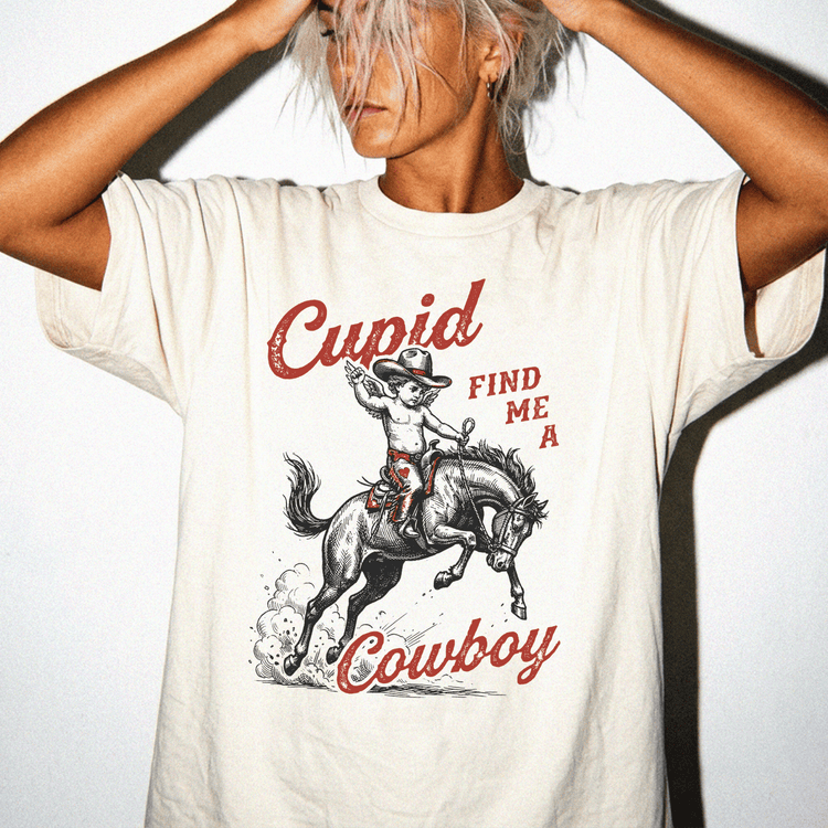 A woman wearing a white t-shirt with a graphic design of a cowboy riding a horse and the text "Cupid FIND ME A COWBOY" printed on it.