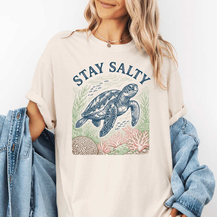 A woman wearing a white t-shirt with a graphic design of a sea turtle and coral reef, along with the text "STAY SALTY".