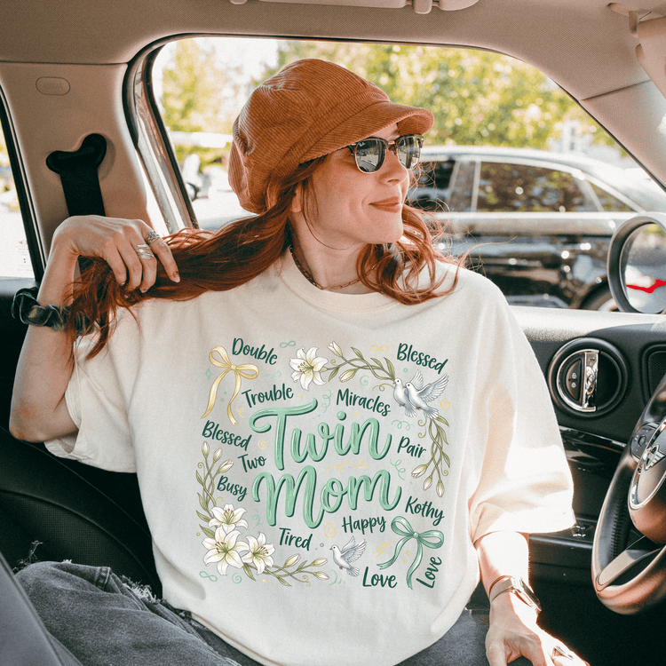 A woman wearing a white t-shirt with a floral design and the words "Twin Mom" is sitting in a car, smiling and looking out the window.