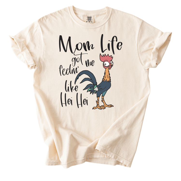A white t-shirt with a graphic design featuring a cartoon rooster and the text "Mom Life got me feelin' like Hei Hei" printed on it.
