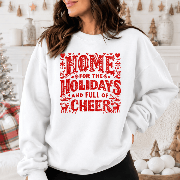 A woman wearing a white sweatshirt with a red and white design that says "HOME FOR THE HOLIDAYS AND FULL OF CHEER".