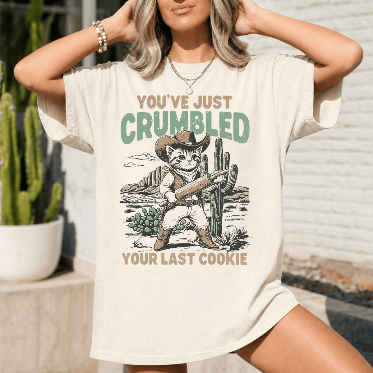 A woman wearing a white t-shirt with a graphic design of a cat holding a gun and the text "You've just Crumbled Your Last Cookie" printed on it.