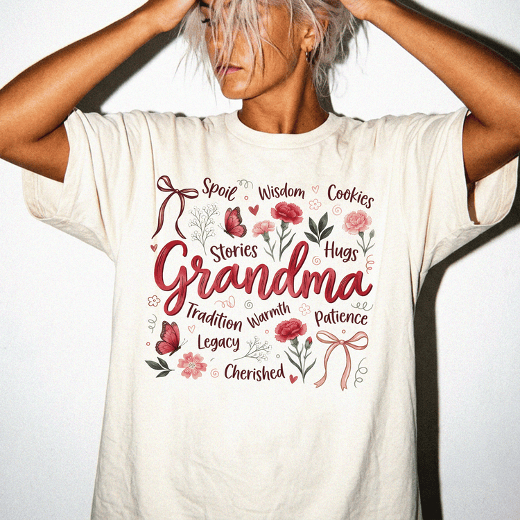 A woman wearing a white t-shirt with a graphic design featuring the word "Grandma" surrounded by various words and illustrations such as "Spoil", "Wisdom", "Cookies", "Hugs", "Stories", "Legacy", and "Charmed".