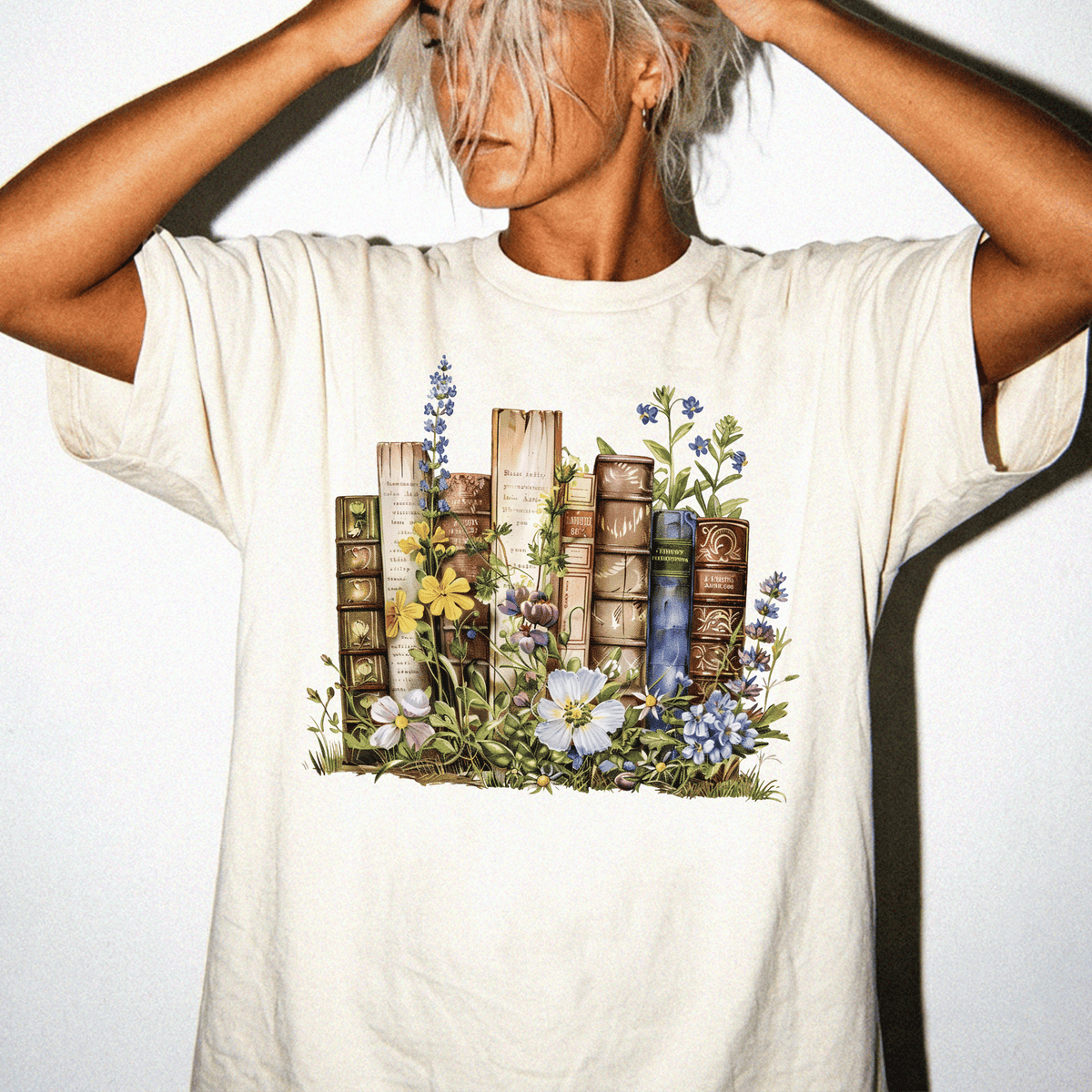A person wearing a white t-shirt with a printed design of books surrounded by flowers and plants.