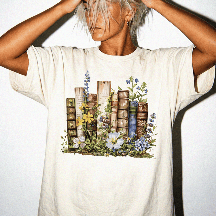 A person wearing a white t-shirt with a printed design of books surrounded by flowers and plants.