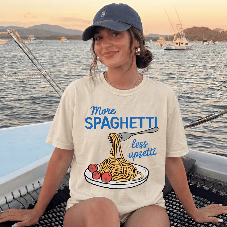 A woman wearing a white t-shirt with a graphic of spaghetti and a fork, sitting on a boat with a scenic background of water and boats.