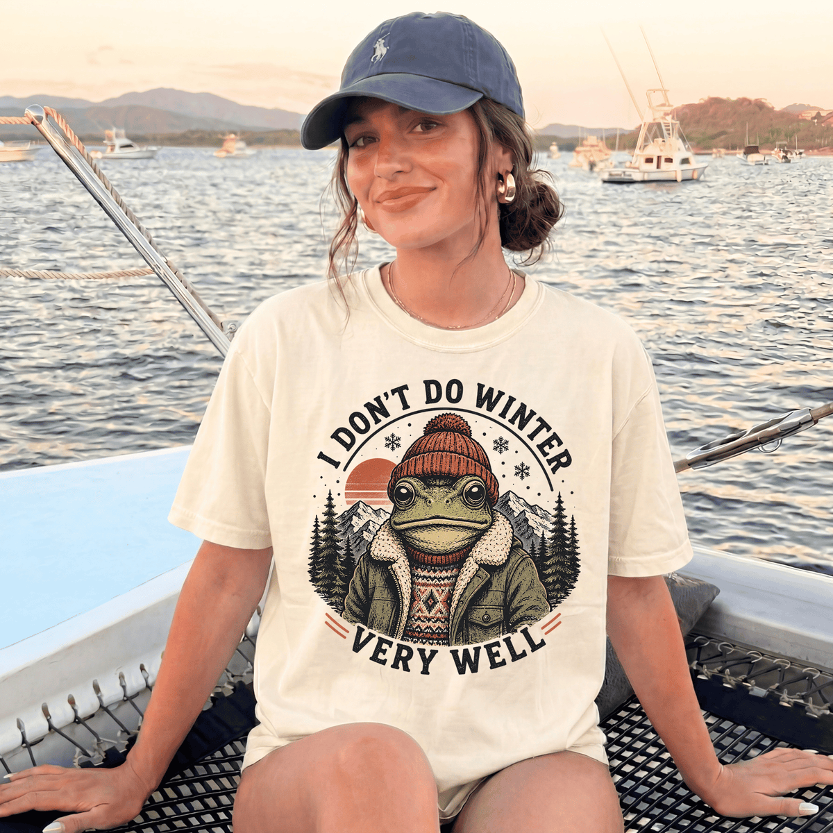 A woman wearing a white t-shirt with a graphic of a frog and the text "I DON'T DO WINTER VERY WELL" is sitting on a boat in the water.