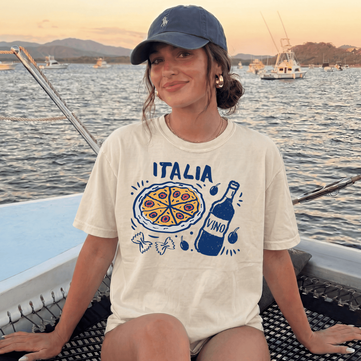 A woman wearing a cap and a t-shirt with a graphic of pizza and wine is sitting on a boat in the water.