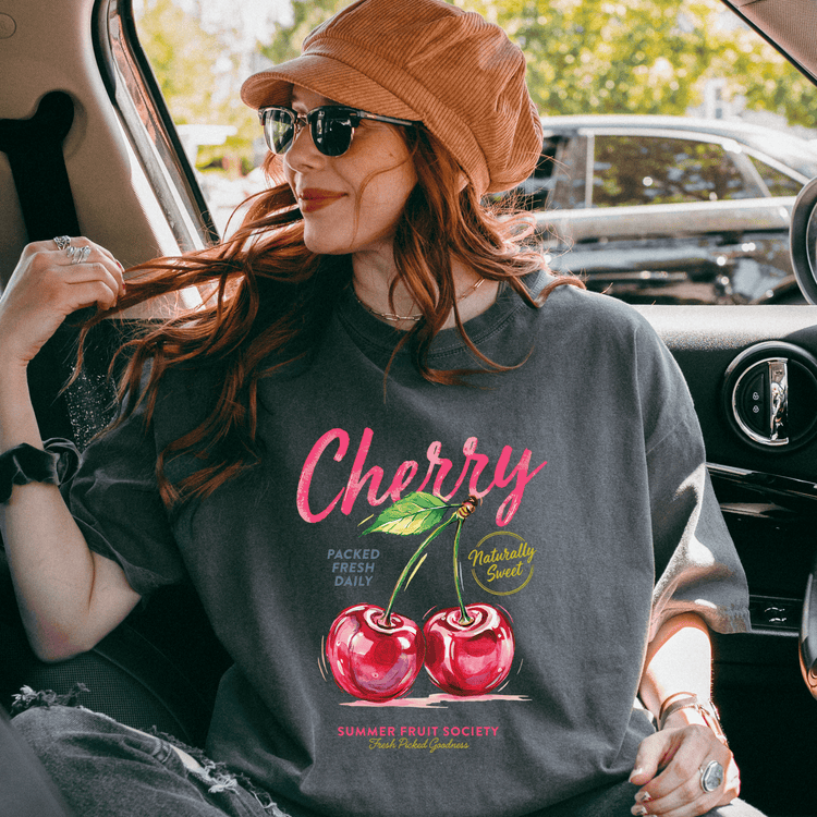 A woman wearing a hat and sunglasses is sitting in a car, looking out the window. She is wearing a gray t-shirt with a graphic design of cherries and text that says "cherry" and "packed fresh daily".