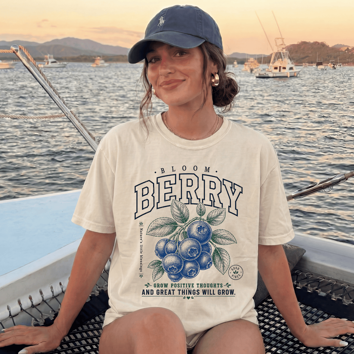 A woman wearing a white t-shirt with a graphic of blueberries and the text "Bloom Berry" is sitting on a boat in the water.