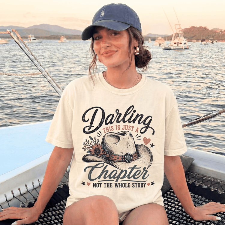 A woman wearing a hat and a t-shirt with a cowboy hat design is sitting on a boat in the water.