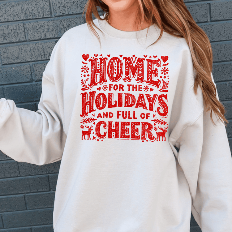 A white sweatshirt with a red and black text design that says "HOME FOR THE HOLIDAYS AND FULL OF CHEER".