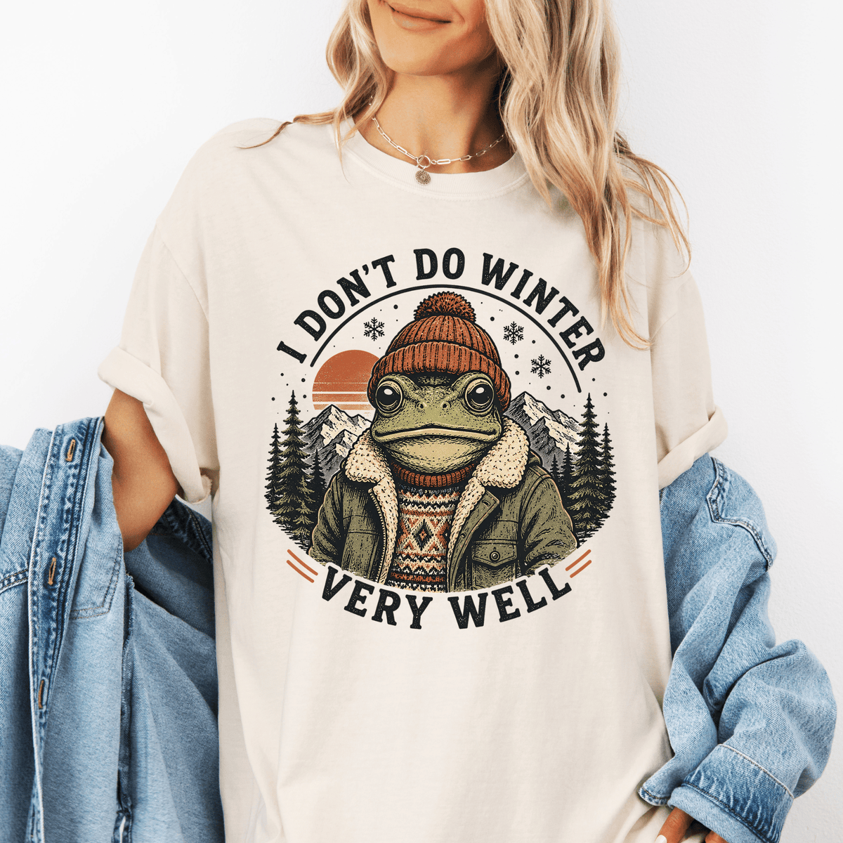 A woman wearing a white t-shirt with a graphic design of a frog wearing winter clothing and a hat, surrounded by snowflakes and mountains.