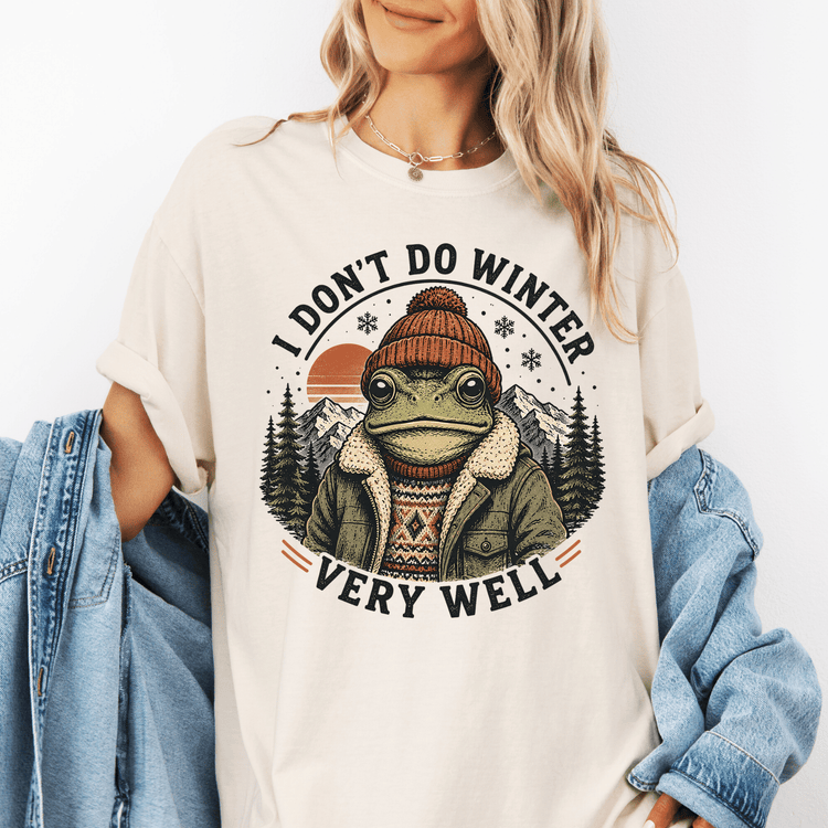 A woman wearing a white t-shirt with a graphic design of a frog wearing winter clothing and a hat, surrounded by snowflakes and mountains.