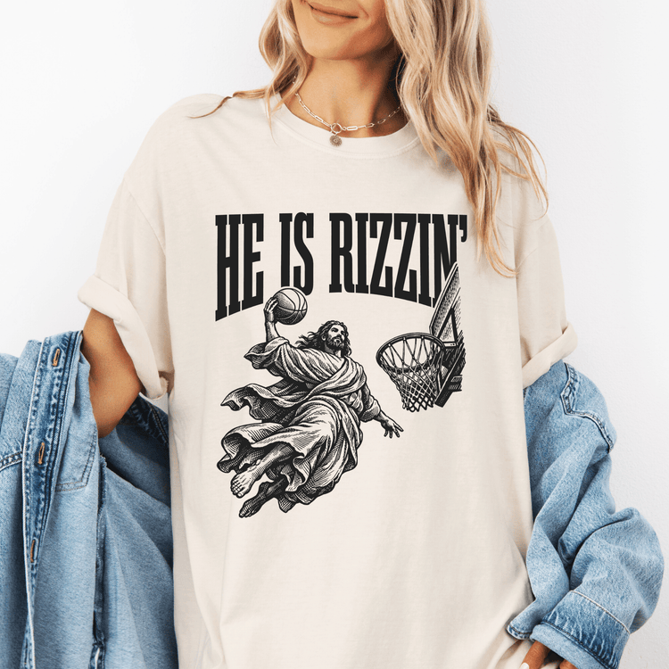 A woman wearing a white t-shirt with a graphic design depicting a basketball player in mid-air, with the text "HE IS RIZZZIN'" printed on it.