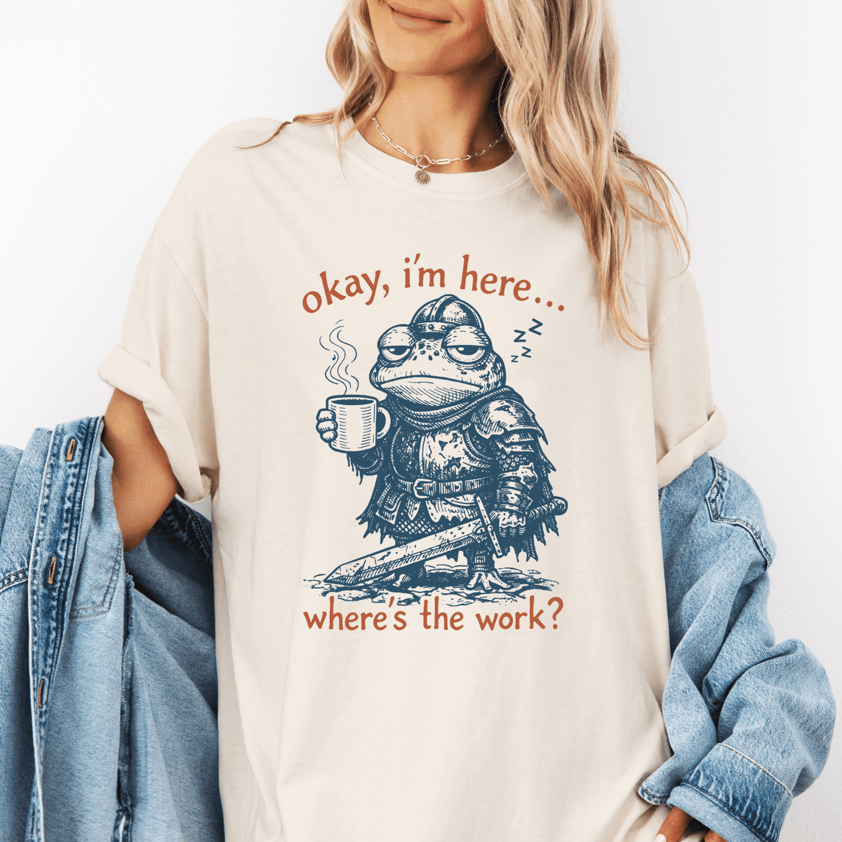 A woman wearing a white t-shirt with a graphic design of a frog holding a cup and posing with the text "okay, im here... where's the work?"