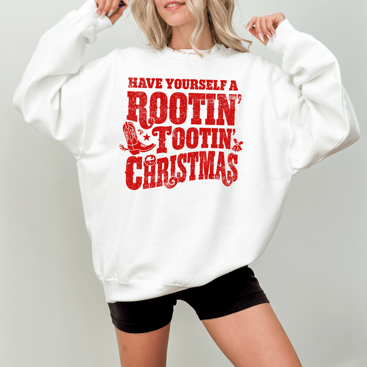 A woman wearing a white sweatshirt with a red text design that says "Have Yourself a Rootin' Tootin' Christmas".