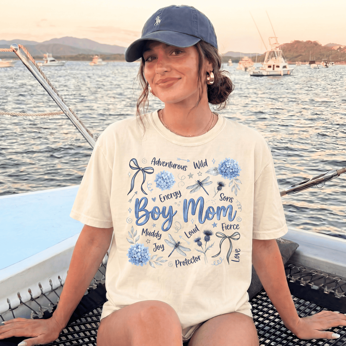 A woman wearing a white t-shirt with a graphic design and text, sitting on a boat in a body of water with other boats in the background.