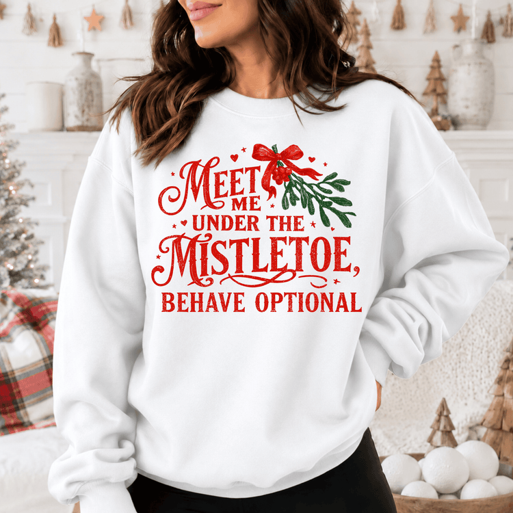 A woman wearing a white sweatshirt with a red and green text design that says "Meet me under the mistletoe, behave optional".