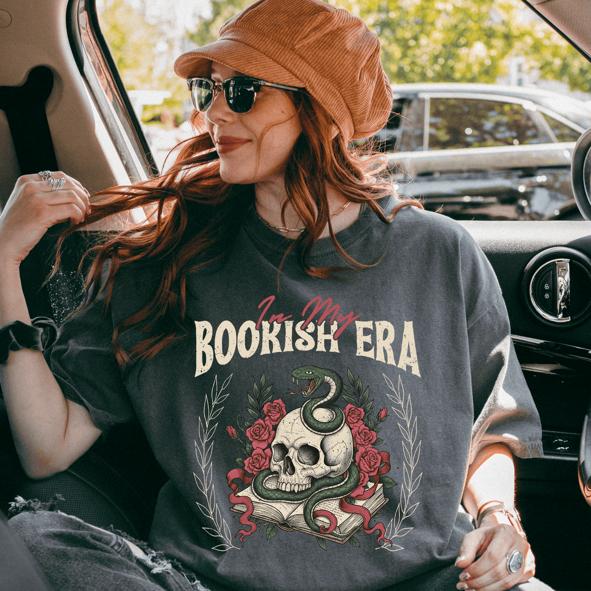 A woman wearing a hat and sunglasses is sitting in a car, wearing a gray t-shirt with a graphic design featuring a skull and a snake.