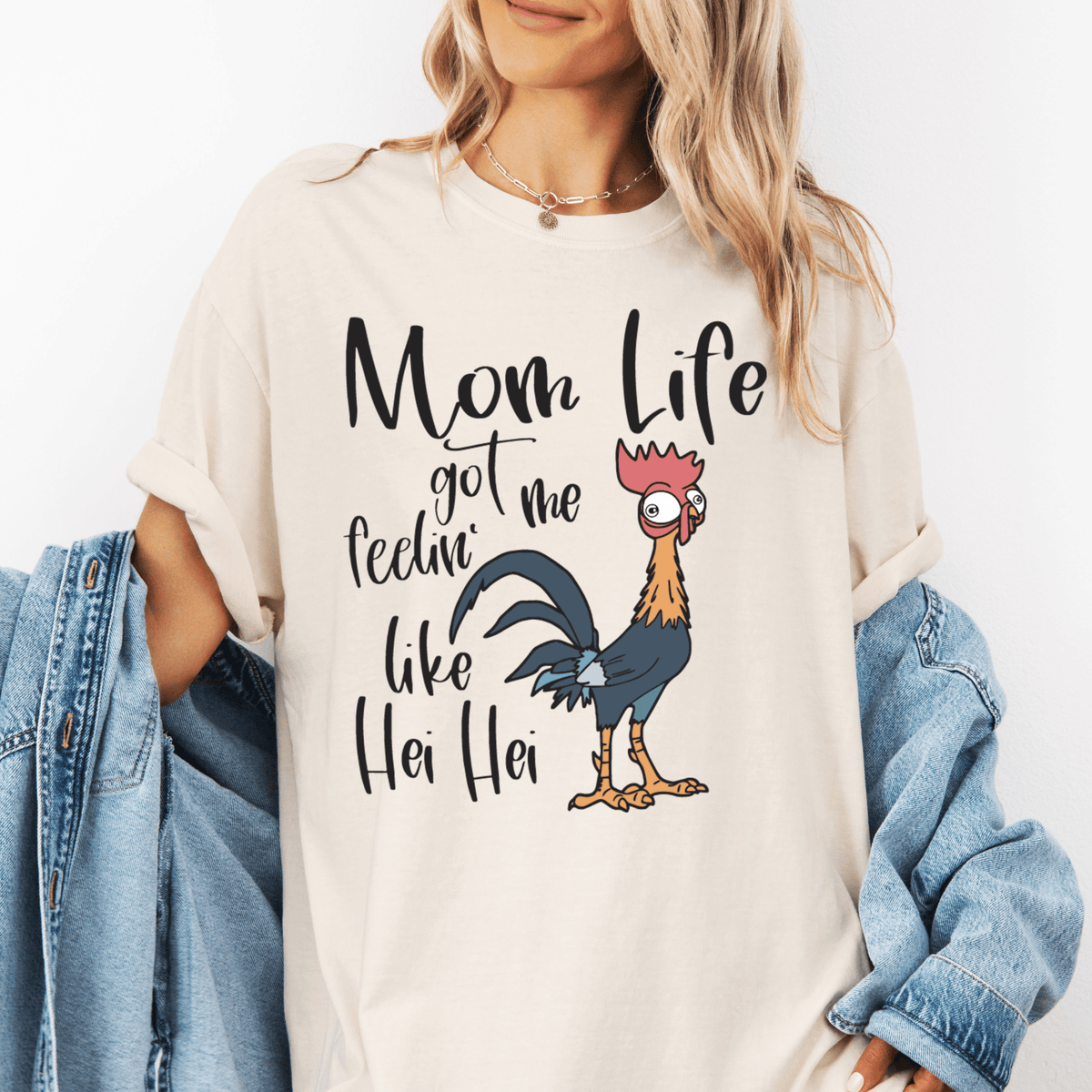 A woman wearing a white t-shirt with a graphic design of a rooster and the text "Mom Life got me feelin' like Hei Hei" printed on it.