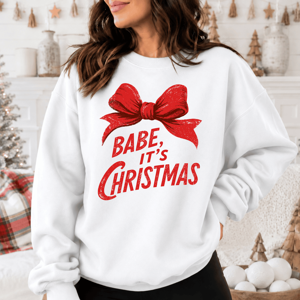 A woman wearing a white sweatshirt with a red bow and the text "BABE, IT'S CHRISTMAS" printed on it.