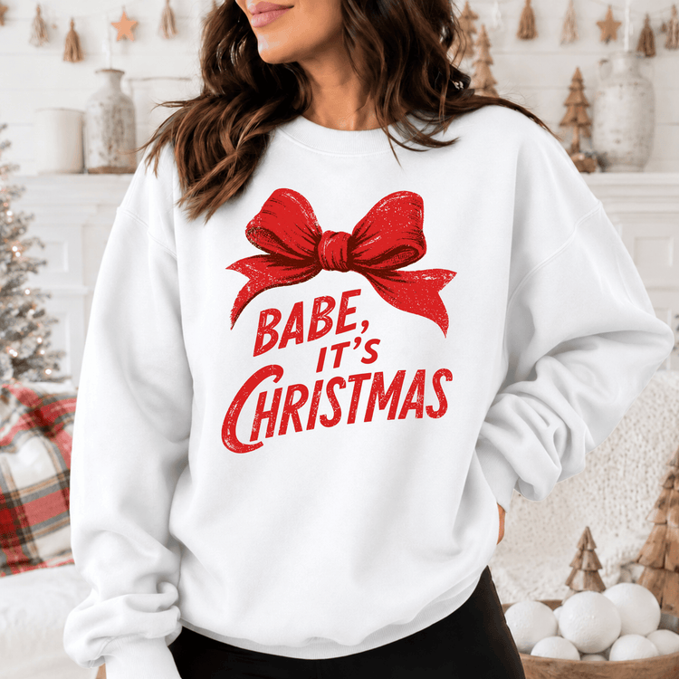 A woman wearing a white sweatshirt with a red bow and the text "BABE, IT'S CHRISTMAS" printed on it.