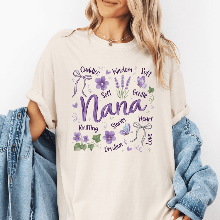 A woman wearing a white t-shirt with a graphic design featuring the word "Nana" surrounded by various purple elements, such as flowers, ribbons, and butterflies.