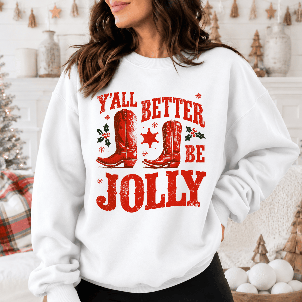 A woman wearing a white sweatshirt with a red graphic design that says "Y'ALL BETTER BE JOLY" and features red cowboy boots and holly leaves.