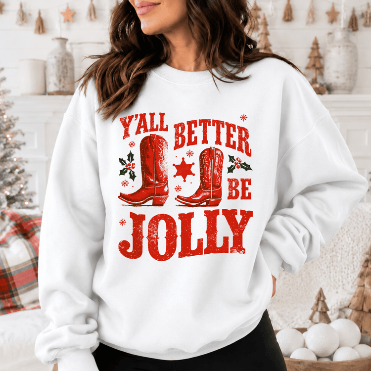 A woman wearing a white sweatshirt with a red graphic design that says "Y'ALL BETTER BE JOLY" and features red cowboy boots and holly leaves.