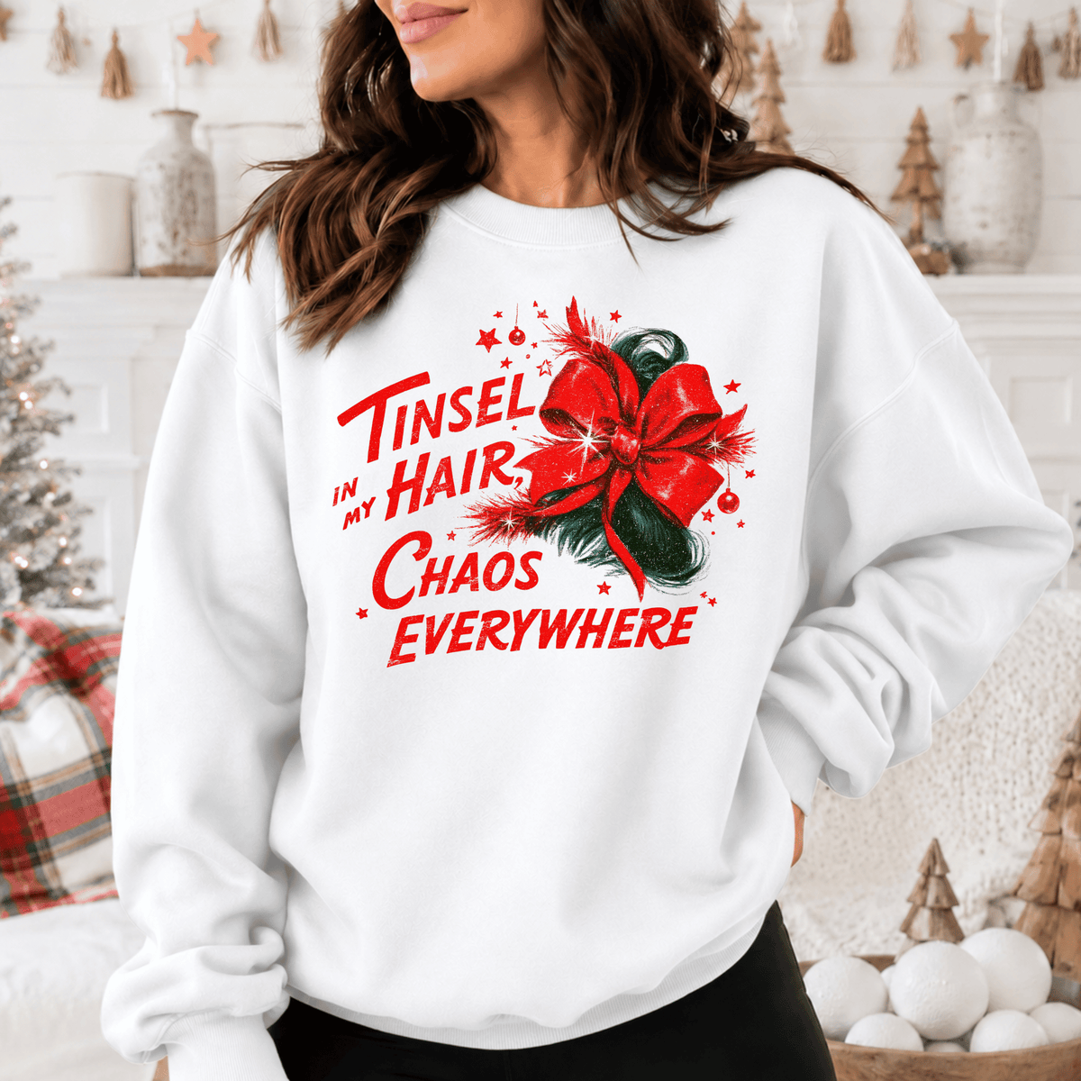 A woman wearing a white sweatshirt with a red bow and text printed on it, standing in front of a Christmas tree and other holiday decorations.