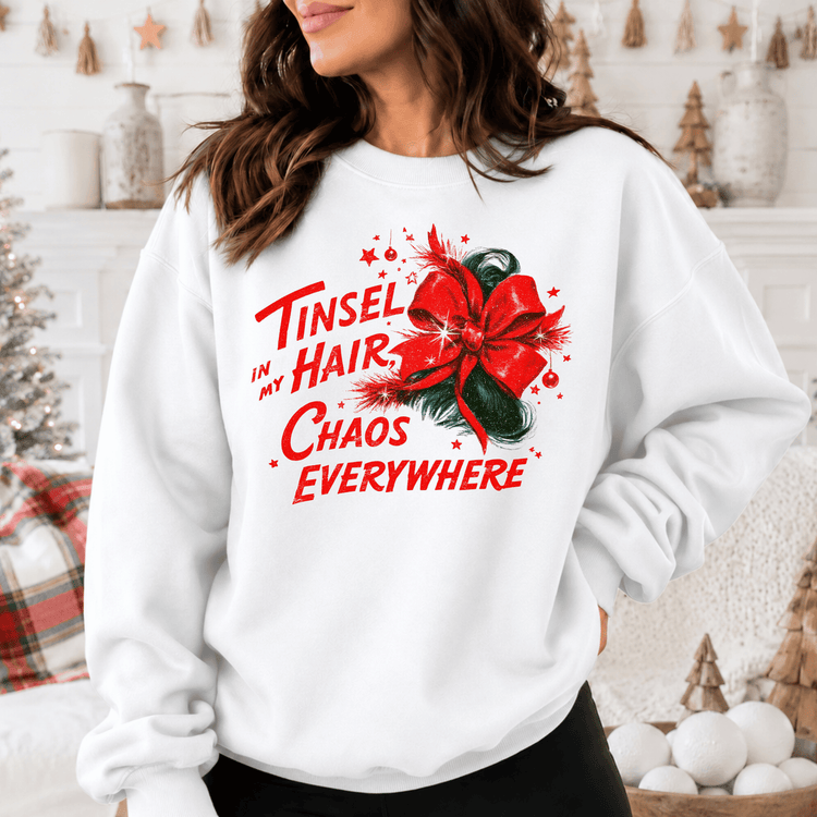 A woman wearing a white sweatshirt with a red bow and text printed on it, standing in front of a Christmas tree and other holiday decorations.