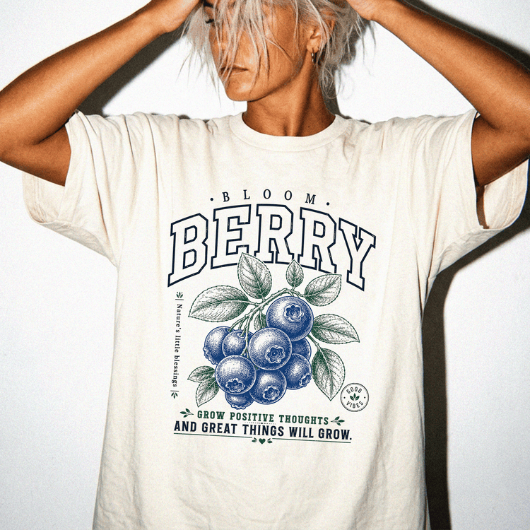 A woman wearing a white t-shirt with a graphic design of blueberries and leaves, along with the text "Bloom Berry" and a motivational quote.