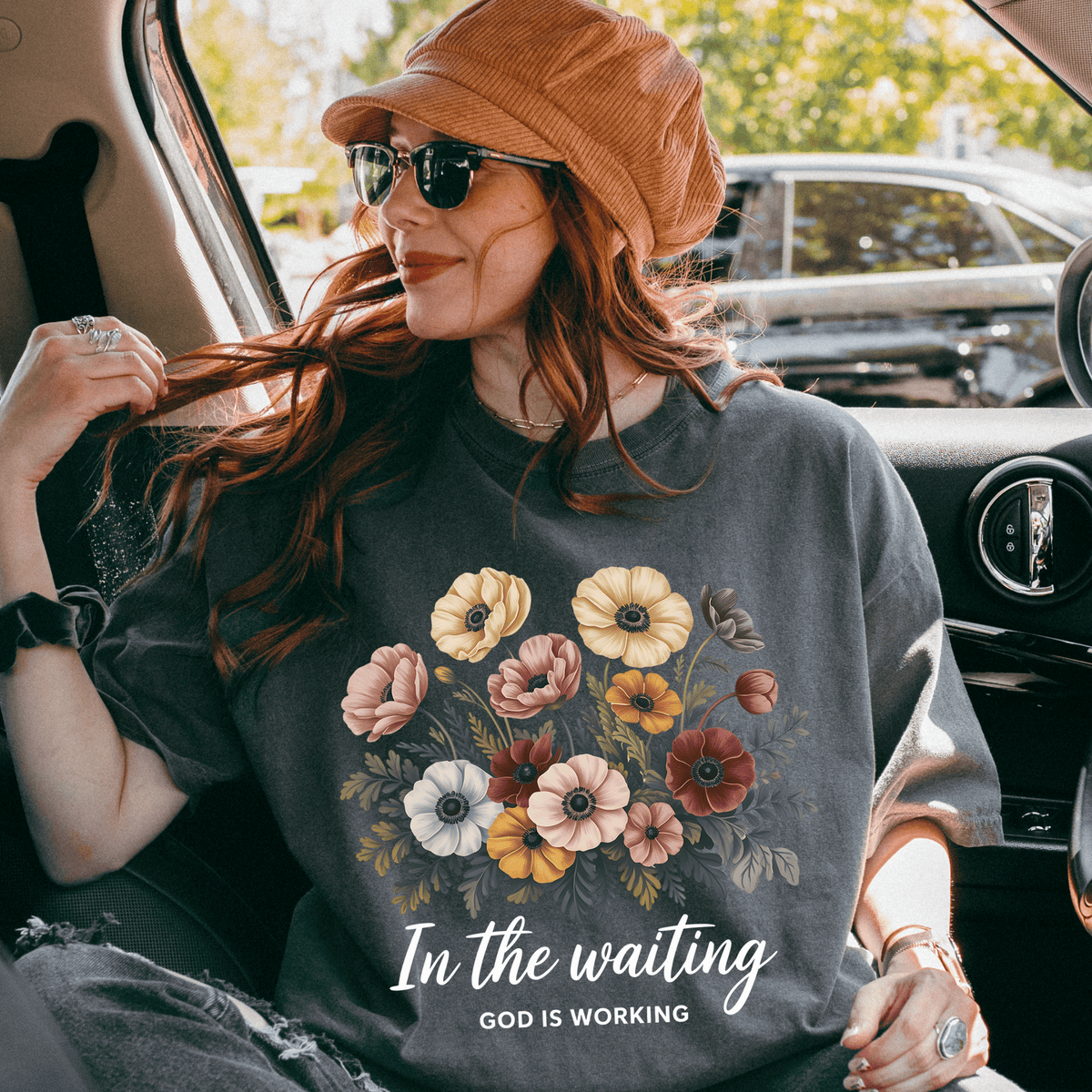 A woman wearing a floral t-shirt and a hat is sitting in the back seat of a car, smiling and looking out the window.