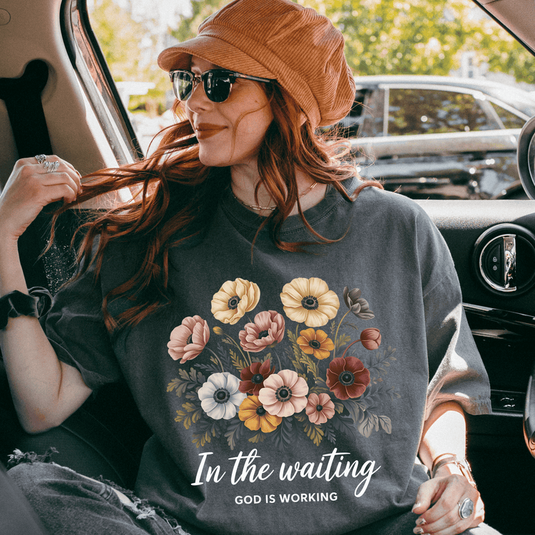 A woman wearing a floral t-shirt and a hat is sitting in the back seat of a car, smiling and looking out the window.