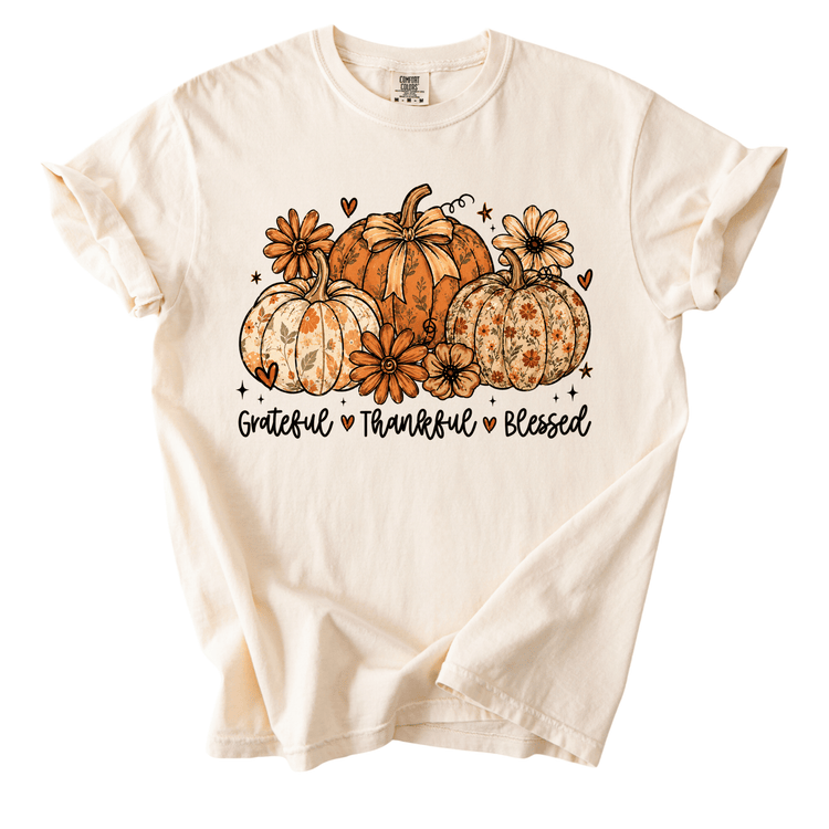 A white t-shirt with a graphic design featuring three pumpkins and flowers, along with the text "Grateful, Thankful, Blessed" printed on it.