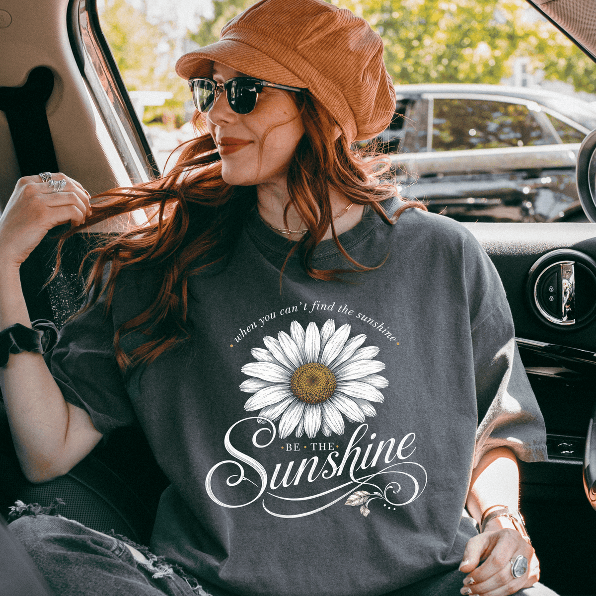 A woman wearing a gray t-shirt with a white daisy flower design and the text "Be the sunshine" is sitting in the back seat of a car.