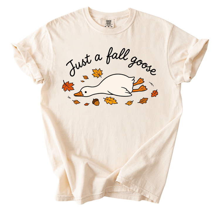 A white t-shirt with a graphic design depicting a duck surrounded by autumn leaves and the text "Just a fall goose" printed on it.